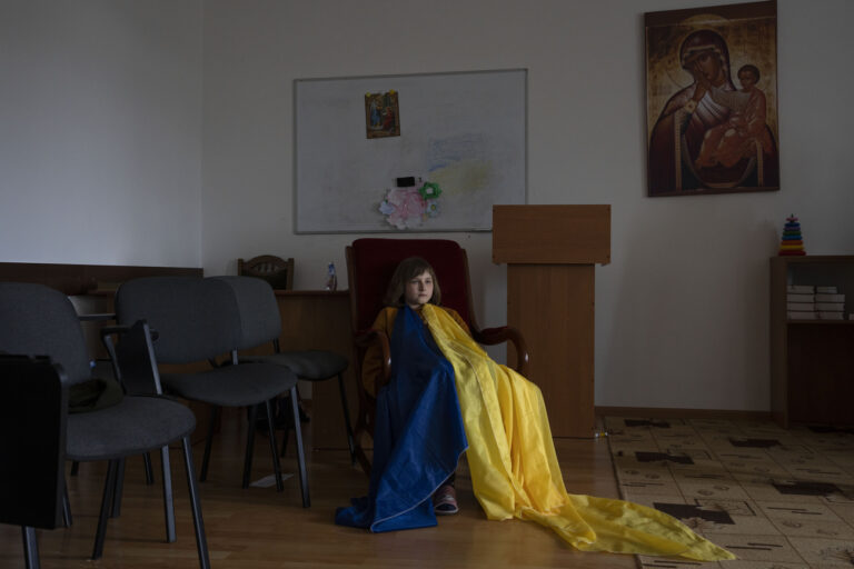 An internally displaced child, wearing a make-shift Ukrainian flag, sits during an activities class with nuns, as she takes refuge at the Hoshiv Women Monastery, in Ivano-Frankivsk region, western Ukraine, Wednesday, April 6, 2022. Beneath the ancient beech forests of the Carpathian Mountains, a quiet monastery in the western Ukrainian village of Hoshiv has transformed itself into a giant playground for a dozen children who've been displaced by the war with their families. (AP Photo/Nariman El-Mofty)