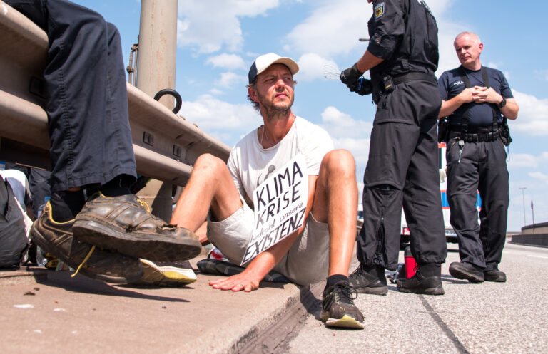 13.08.2022, Hamburg: Ein Klimaaktivist sitzt bei einer Blockade der Köhlbrandbrücke mit seiner an der Brücke festgeklebten Hand am Boden und hält ein Schild mit der Aufschrift 