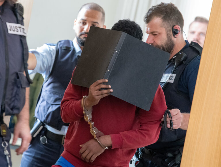 04.07.2023, Baden-Württemberg, Ulm: Der mit Handschellen gefesselte Angeklagte (M), der sein Gesicht mit einem Aktenordner verdeckt, wird von Justizbeamten in den Verhandlungssaal des Landgerichts geführt. Der Asylbewerber aus Eritrea wurde im Fall des Messerangriffs auf zwei Schülerinnen in Illerkirchberg zu einer lebenslangen Freiheitsstrafe verurteilt. Das Gericht sah es als erwiesen an, dass er eine 14-Jährige und ihre 13 Jahre alte Freundin auf dem Schulweg mit einem Messer heimtückisch attackierte. Die 14-Jährige starb an ihren Verletzungen. Foto: Stefan Puchner/dpa +++ dpa-Bildfunk +++ (KEYSTONE/DPA/Stefan Puchner)