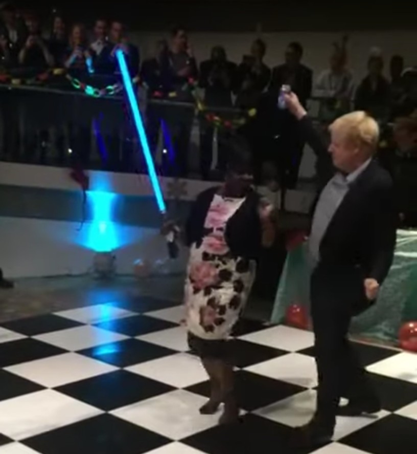 Hier sieht man, wie der britische Premierminister Boris Johnson sich an einer rauschenden Corona-Party ein Tänzchen mit einer Lady liefert. Die Schöne lockt ihn mit einem Licht-Schwert
